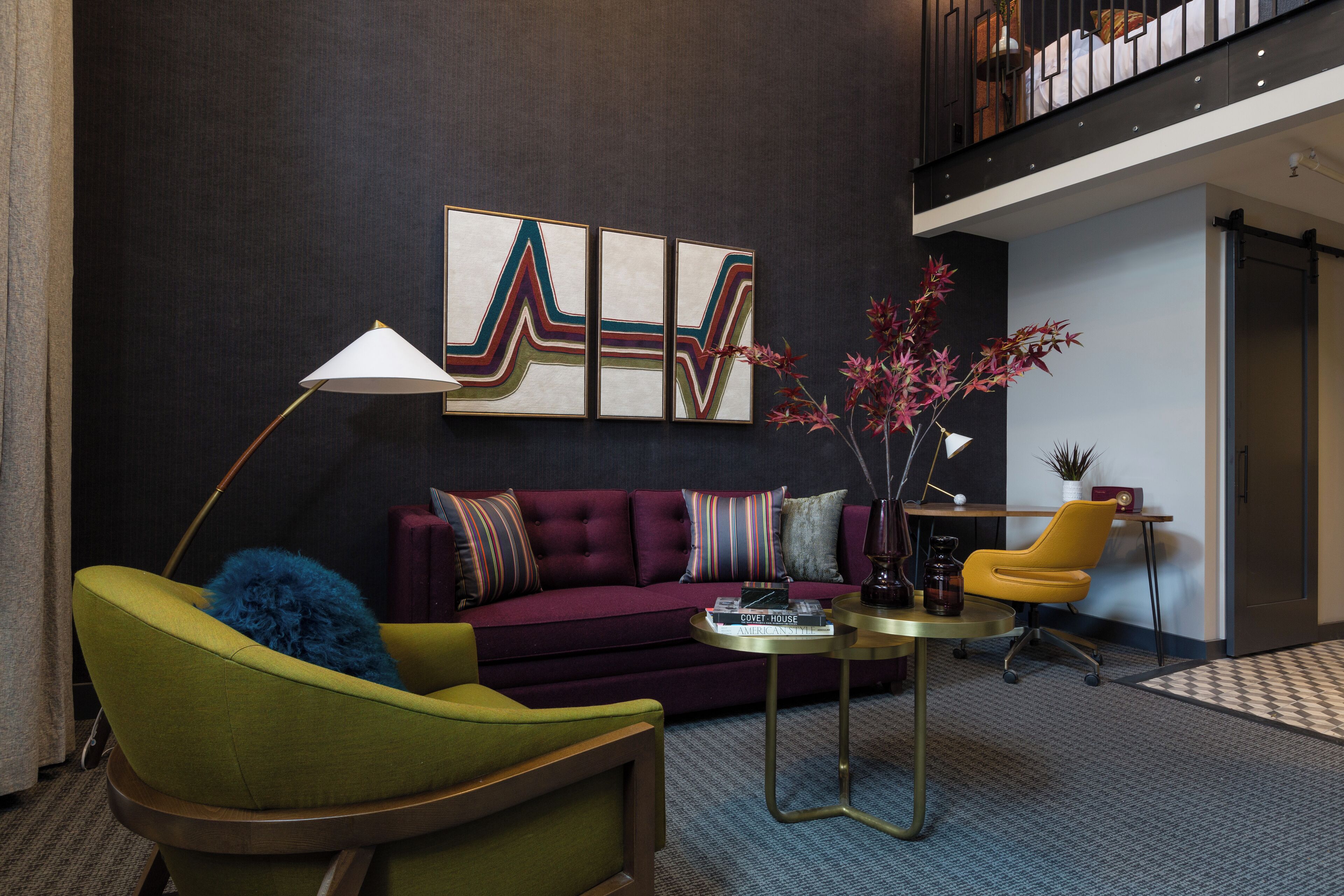 A modern living room with a green armchair, a purple sofa, and a yellow chair. There is a coffee table with books and a lamp. A large abstract painting is on the wall, and a plant is on a side table.