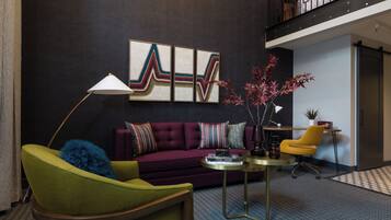 A modern living room with a green armchair, a purple sofa, and a yellow chair. There is a coffee table with books and a lamp. A large abstract painting is on the wall, and a plant is on a side table.