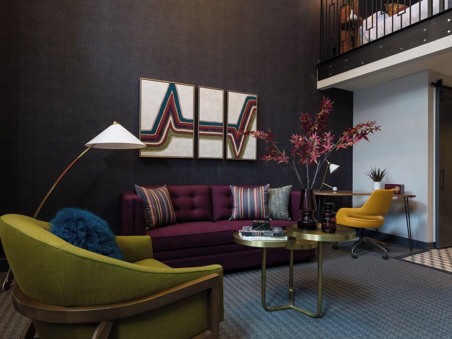 A modern living room with a green armchair, a purple sofa, and a yellow chair. There is a coffee table with books and a lamp. A large abstract painting is on the wall, and a plant is on a side table.