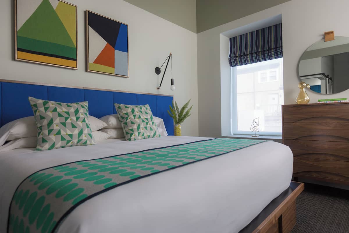 Un lit bien fait, agrémenté d’oreillers à motifs, d’une tête de lit bleue et d’un plaid vert et blanc.