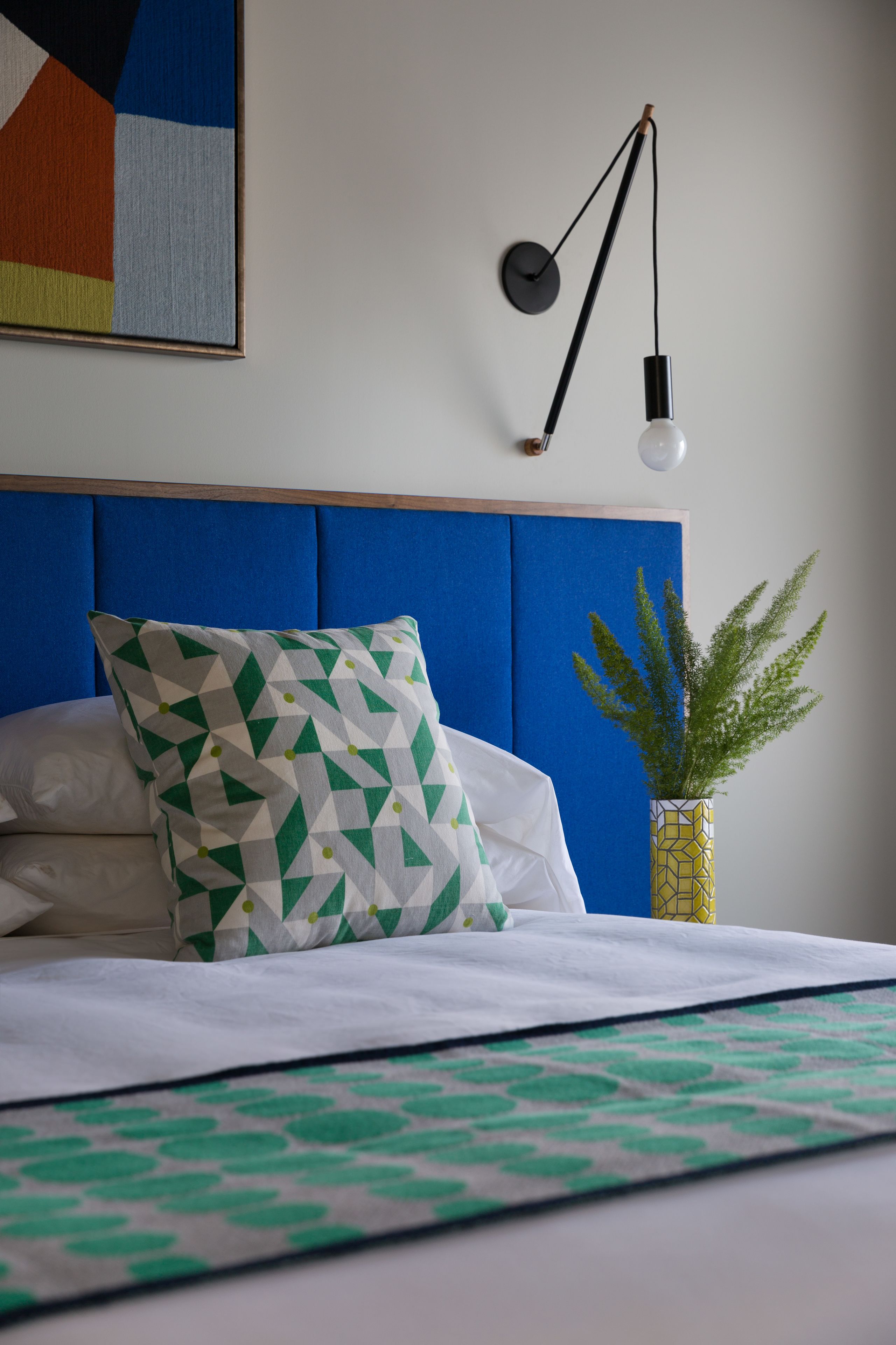 Un lit bien fait, agrémenté d’un coussin aux motifs géométriques, un luminaire fixé au mur et une plante en pot.