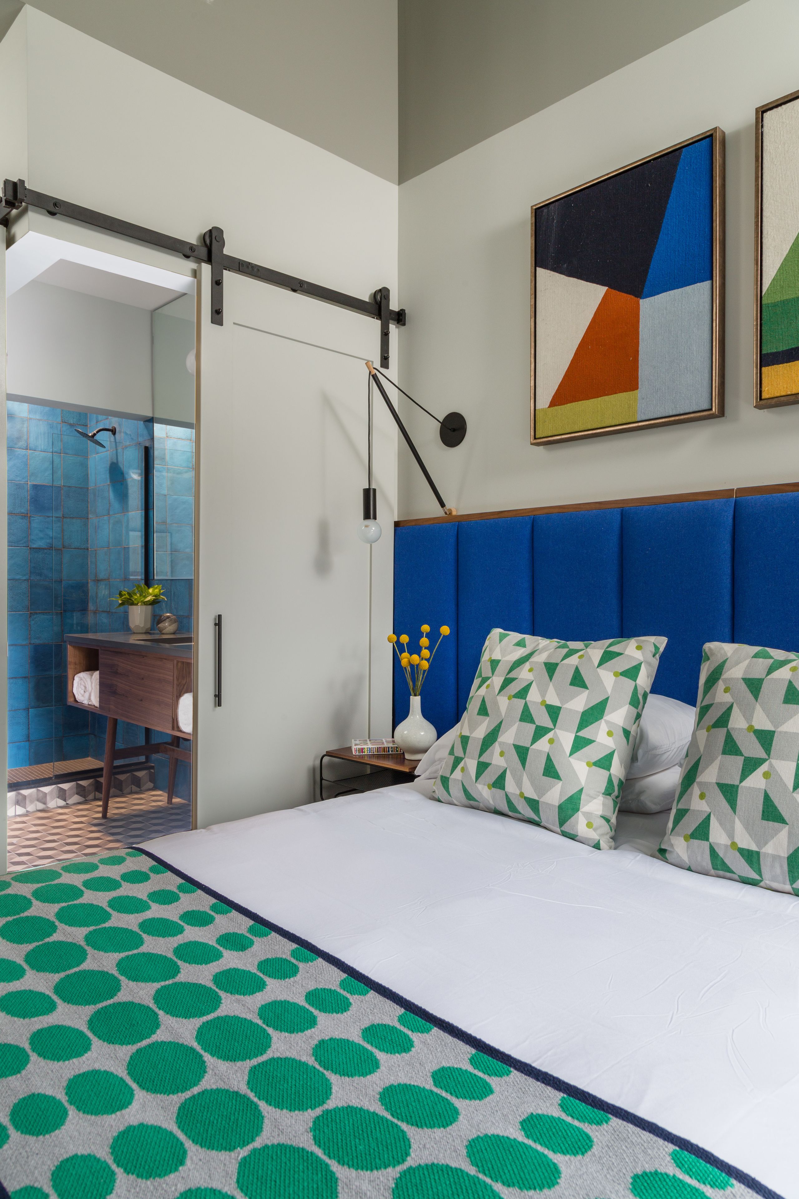 A modern hotel room with a large bed, a blue headboard, a geometric painting, and a view of the bathroom.