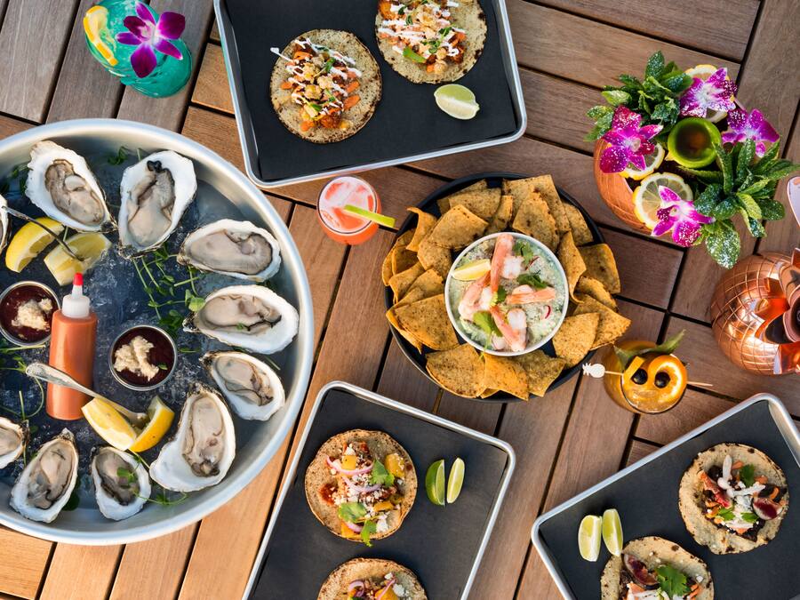 A variety of seafood dishes, including oysters on the half shell, tacos, and a shrimp cocktail, arranged on a wooden table.