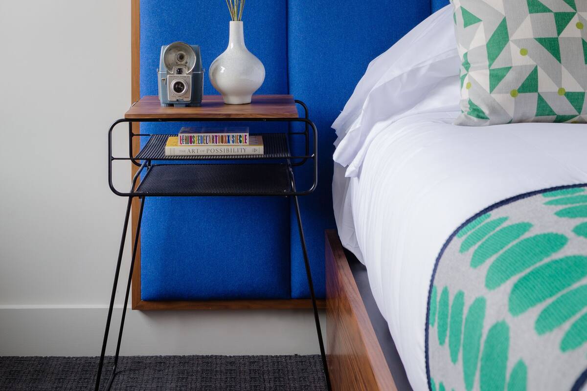 Une chambre à coucher bien rangée, avec une tête de lit bleue, une table de chevet sur laquelle se trouvent un appareil photo et des livres, et un coussin à motifs verts.