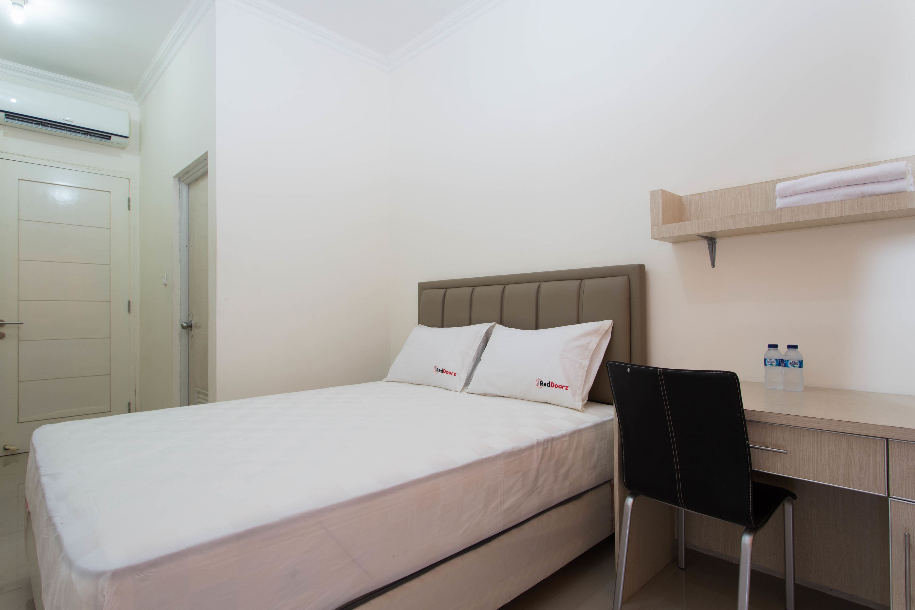 Una cama individual con cabecera, dos almohadas, un escritorio con silla y una estantería con toallas y botellas de agua.