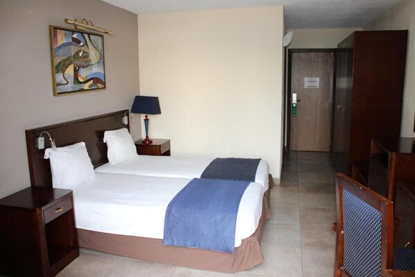 A hotel room with two beds, a nightstand with a lamp, a painting on the wall, and a wardrobe. - Hotel Elais (Pointe-Noire)