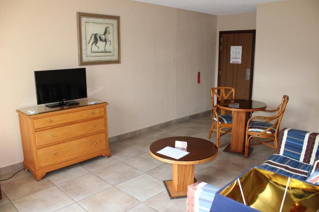 Une pièce comprenant une télévision posée sur une commode en bois, une table ronde avec des chaises et un canapé recouvert d’un tissu à motifs.