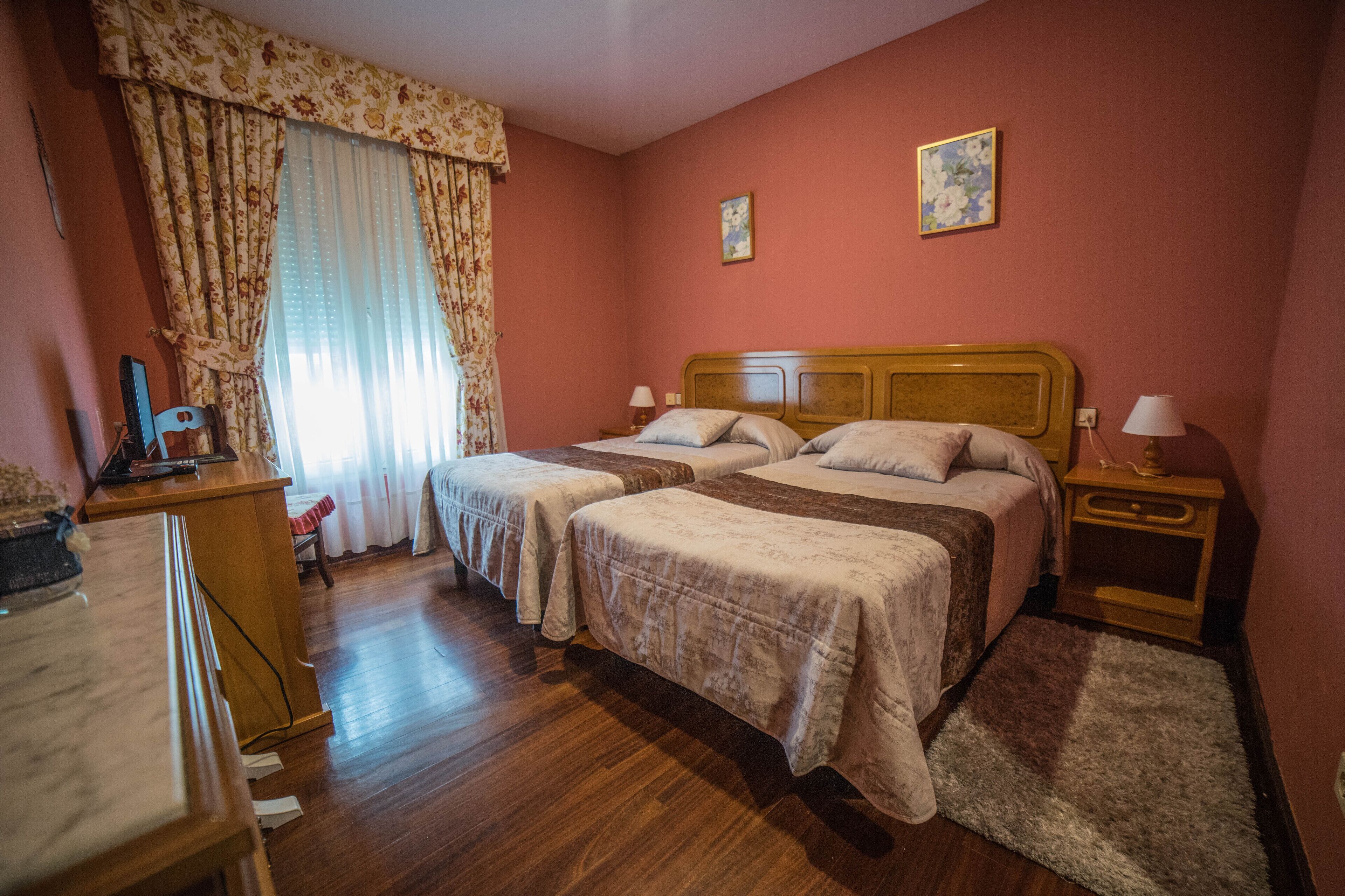 Habitación de hotel con dos camas, un escritorio de madera, una lámpara de escritorio, una mesita de noche y un ventanal con cortinas.