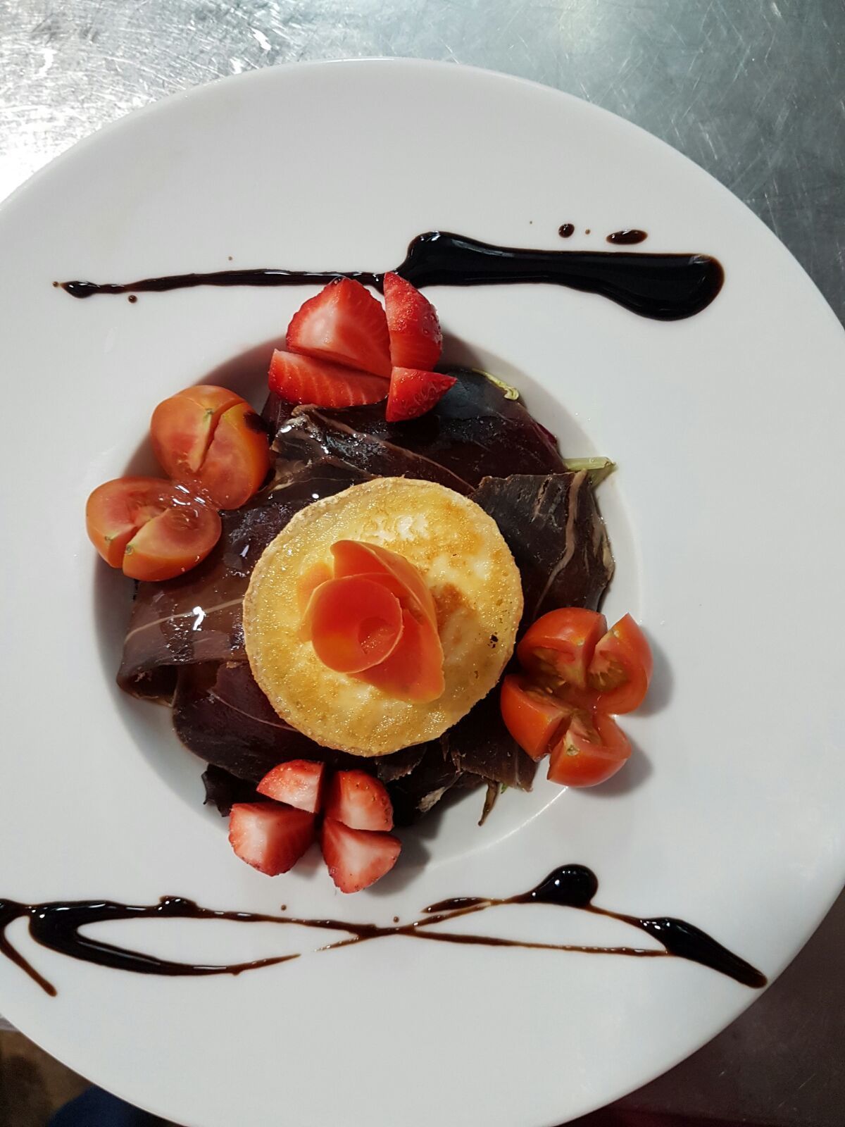 Un plato blanco con un alimento de color oscuro, adornado con fresas y un detalle decorativo, bañado en salsa de chocolate.