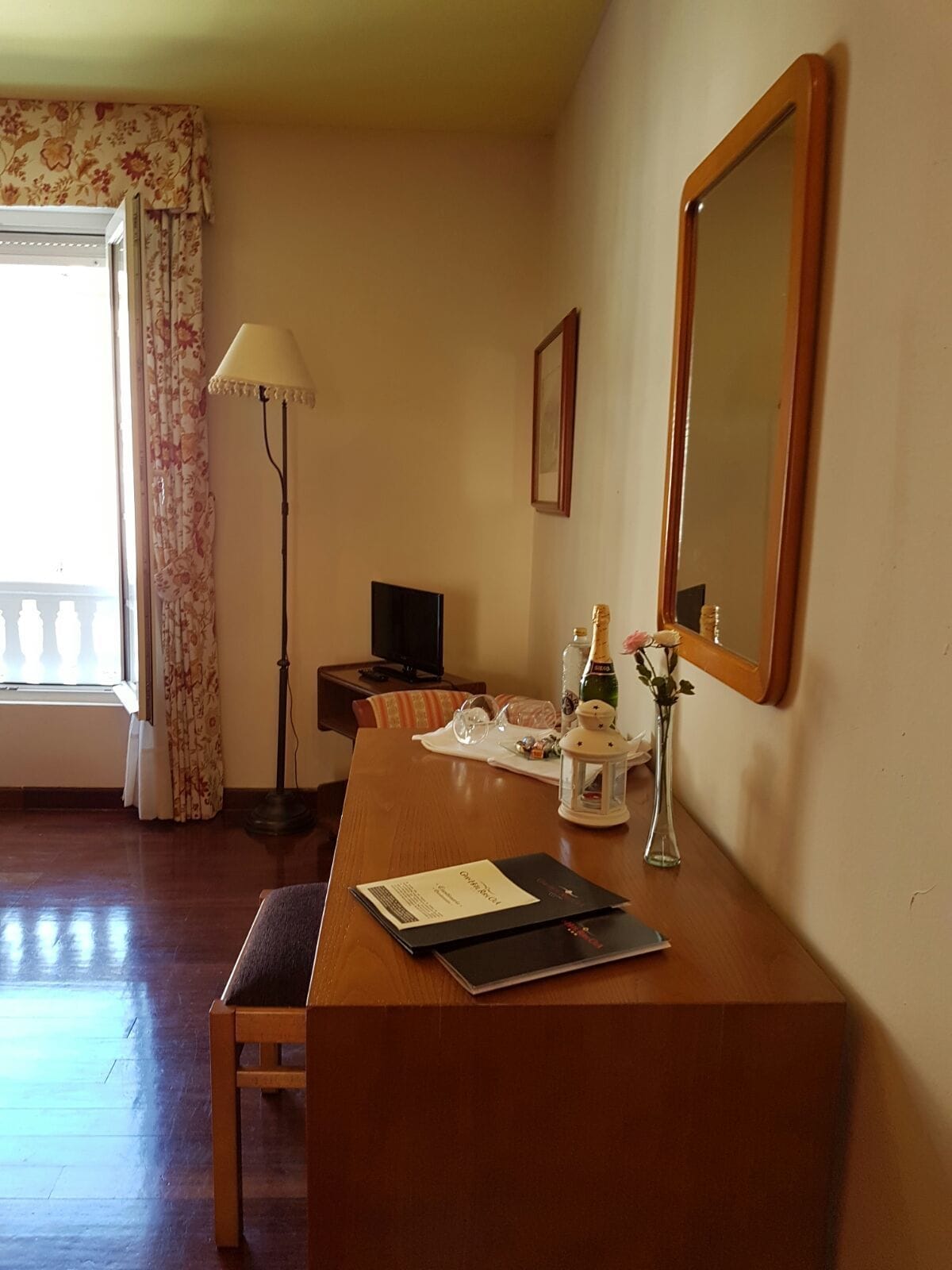 Habitación de hotel con un escritorio de madera, una silla, televisor, lámpara, espejo y ventana con cortinas estampadas.