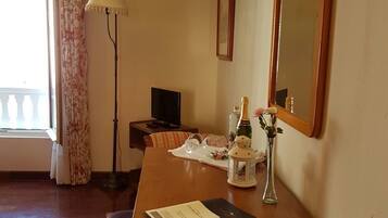 Habitación de hotel con un escritorio de madera, una silla, televisor, lámpara, espejo y ventana con cortinas estampadas.