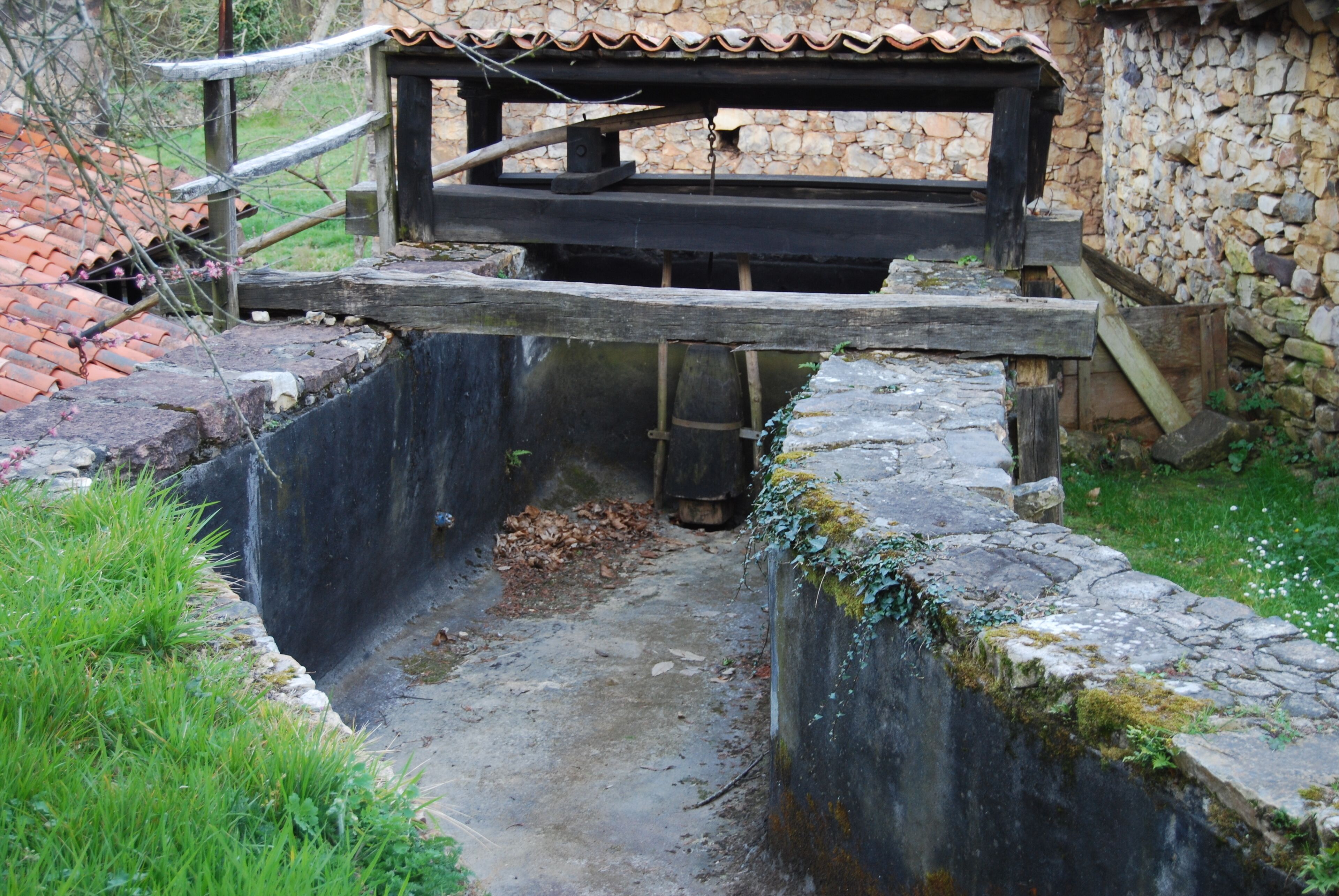 A stone watermill with a wooden structure above it, surrounded by greenery and a stone wall.
