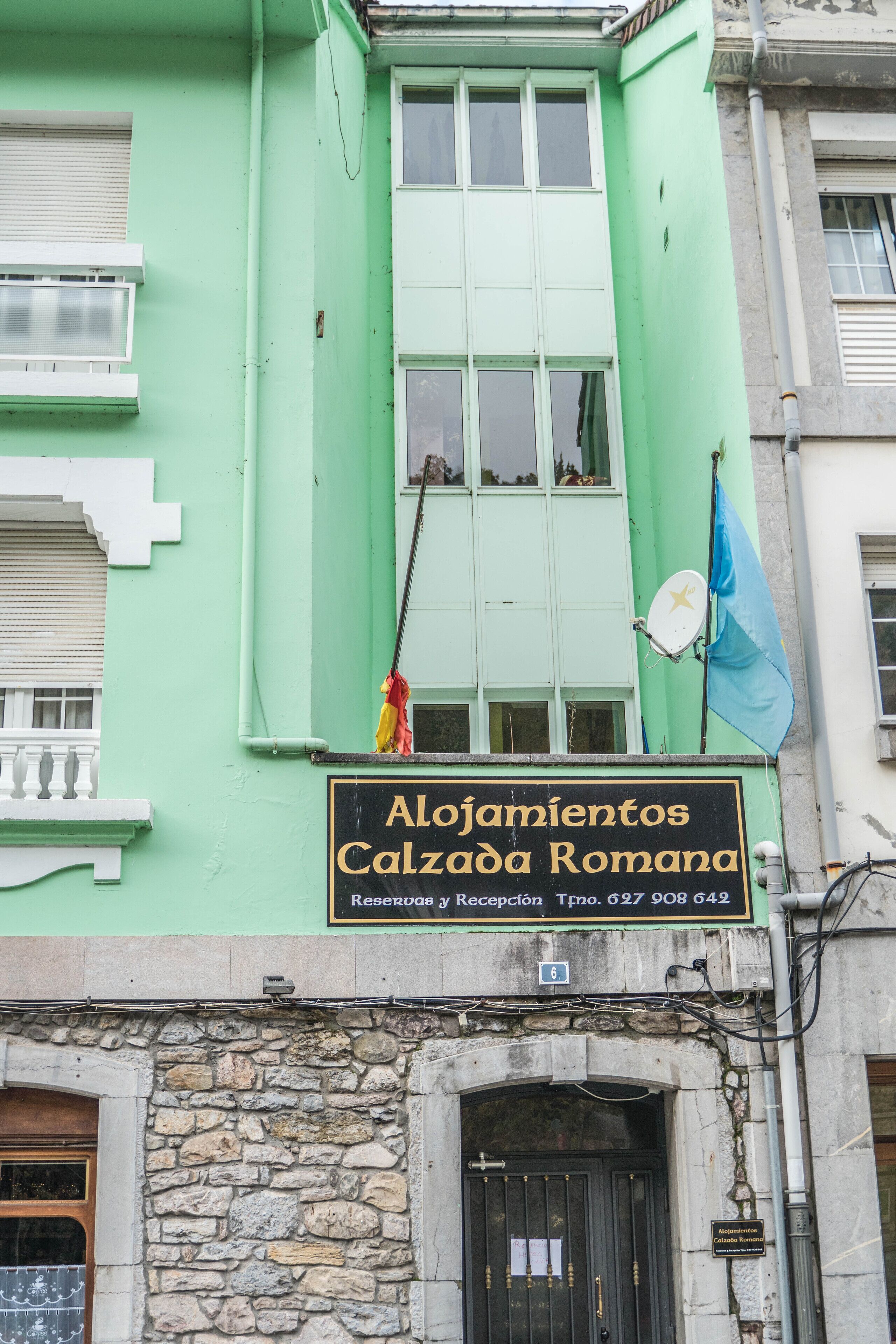 A building with a green facade, a sign reading "Alojamientos Calzada Romana," and a flag.