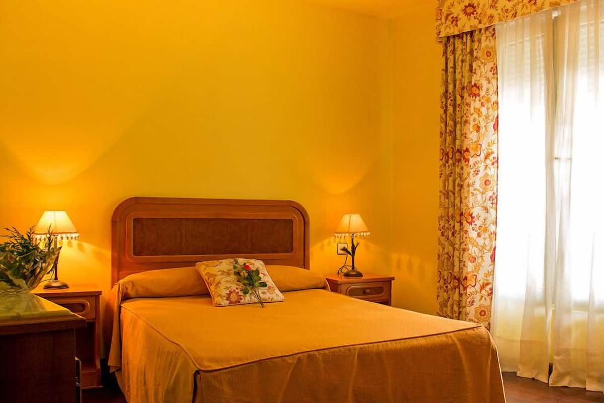 Quarto de hotel com cama, duas luminárias de cabeceira, cortinas florais e janela com cortinas leves.