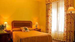 A hotel room with a bed, two bedside lamps, floral curtains, and a window with sheer curtains. - Alojamiento Calzada Romana (Belmonte de Miranda)