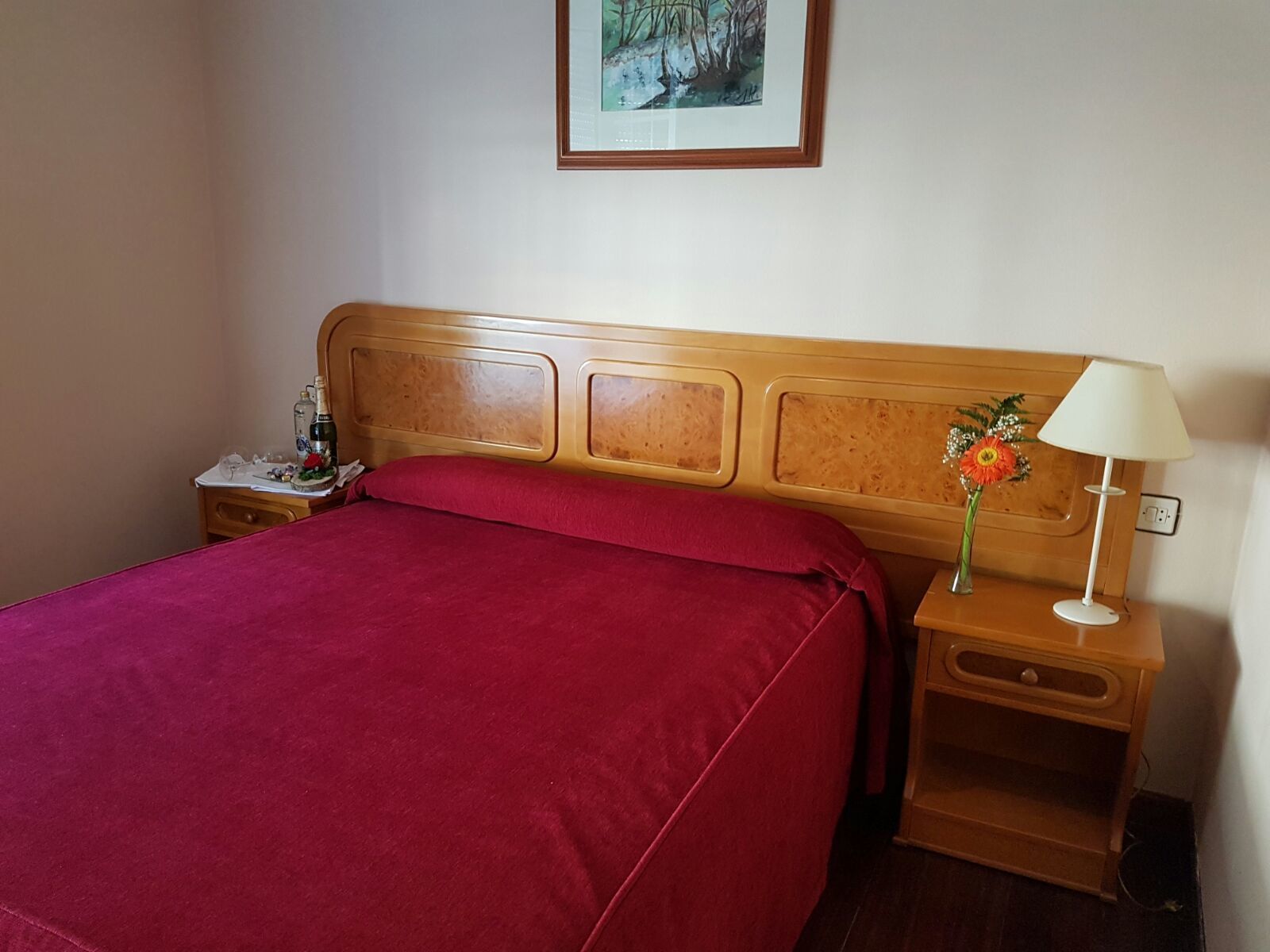 Cama bem arrumada com colcha vermelha e cabeceira de madeira. Apresenta uma mesa de cabeceira com abajur e um vaso de flores. Um quadro emoldurado está pendurado na parede acima da cama.