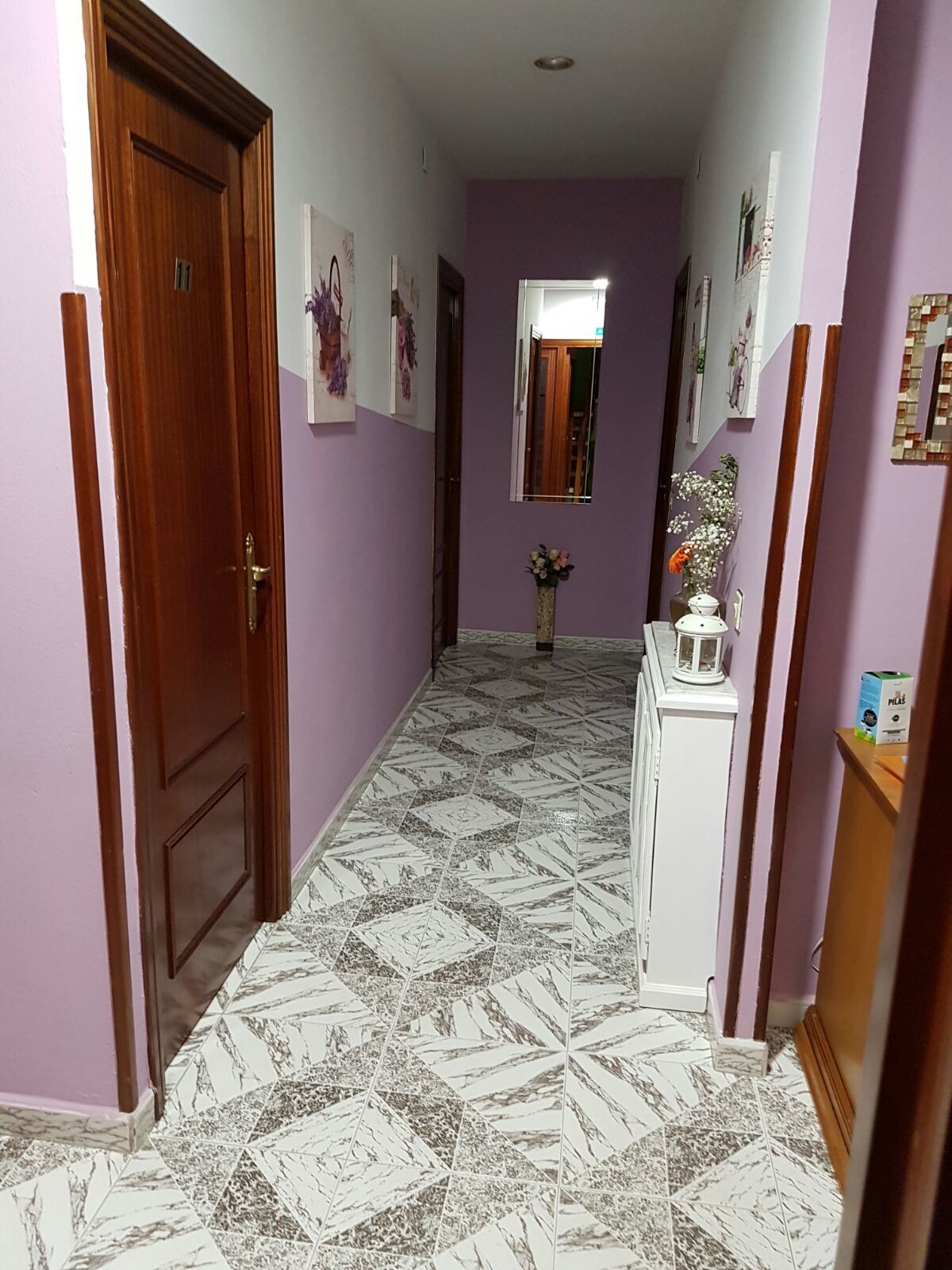 A hallway with patterned floor tiles, a wooden door, and a white cabinet with a mirror and a vase of flowers.