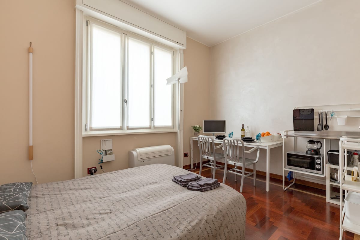 Un dormitorio con cama, escritorio con computadora, silla y kitchenette.