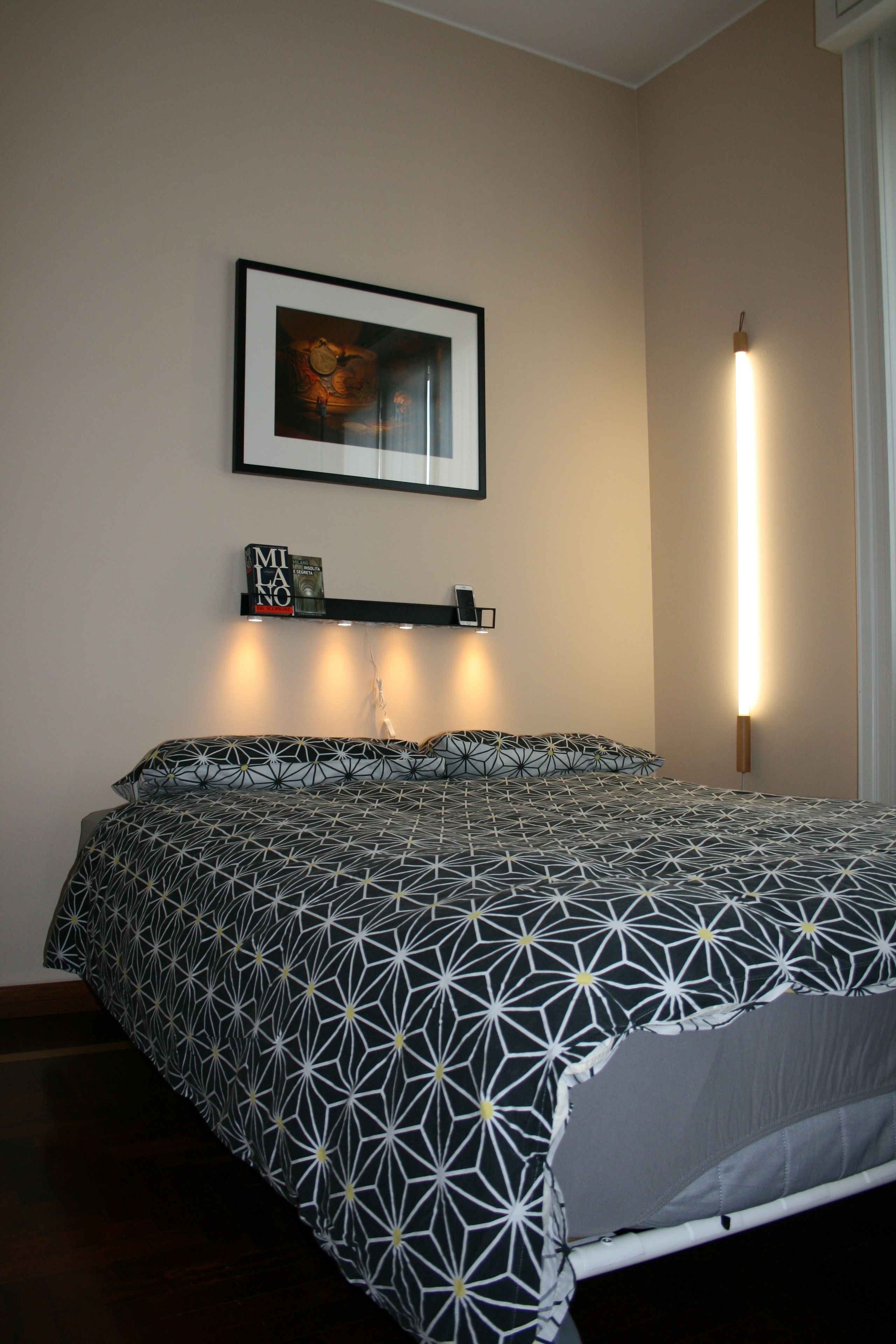 A neatly made bed with a geometric patterned bedspread, a framed artwork above it, and a bedside table with a lamp.