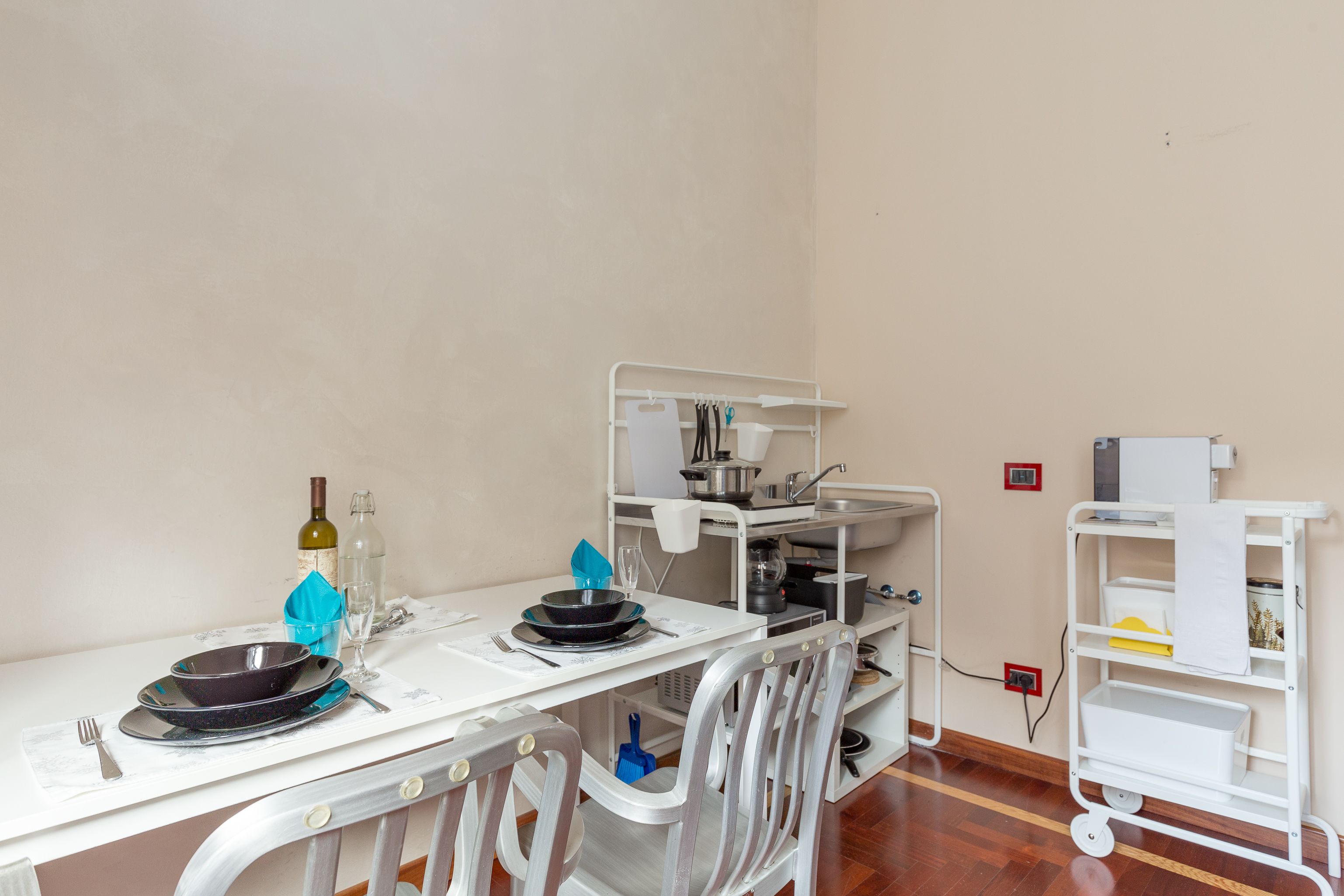 A dining area with a table set for two, a white chair, a white cart with kitchen items, and a wall socket.