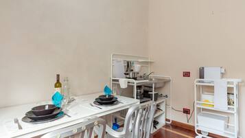 A dining area with a table set for two, a white chair, a white cart with kitchen items, and a wall socket.