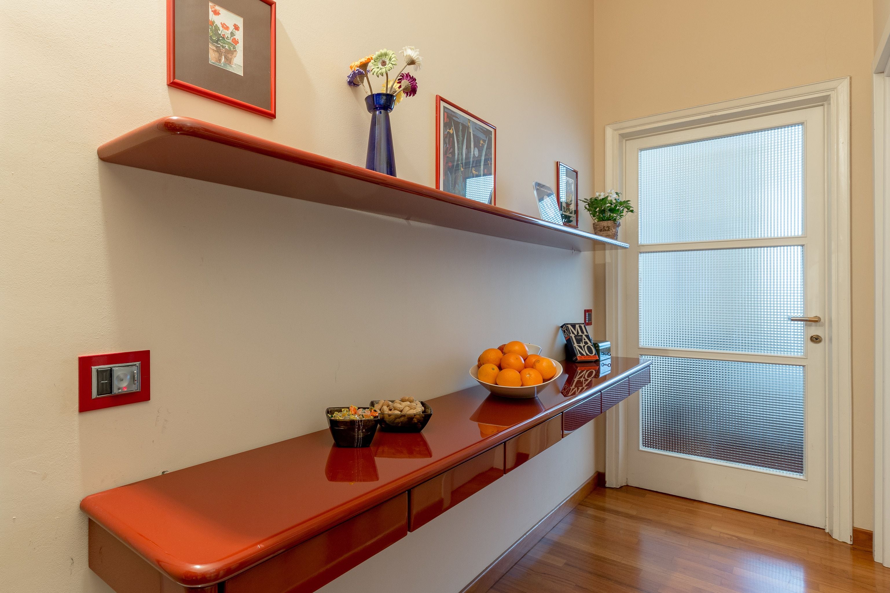 Un couloir avec un banc en bois, un vase rempli de fleurs et un bol d’oranges posé sur une étagère.