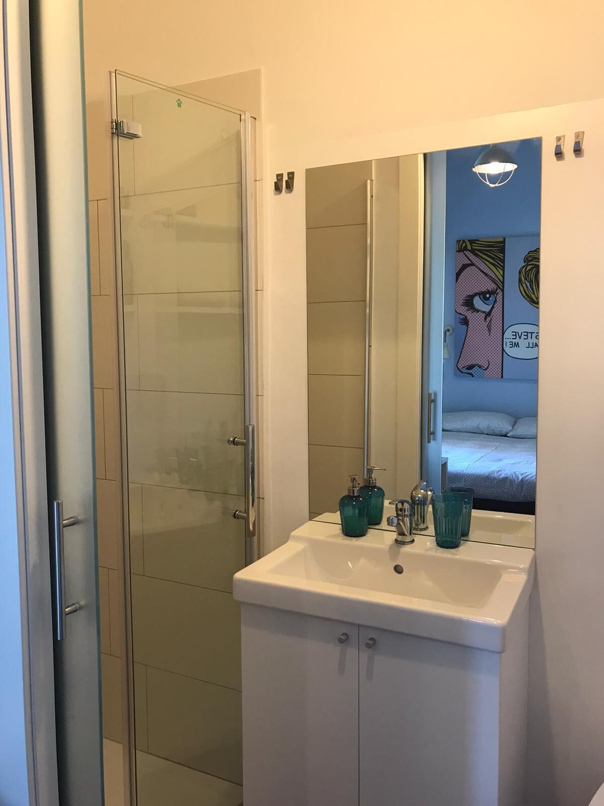 A bathroom with a glass shower door, a white sink with two bottles, and a mirror reflecting a bed.
