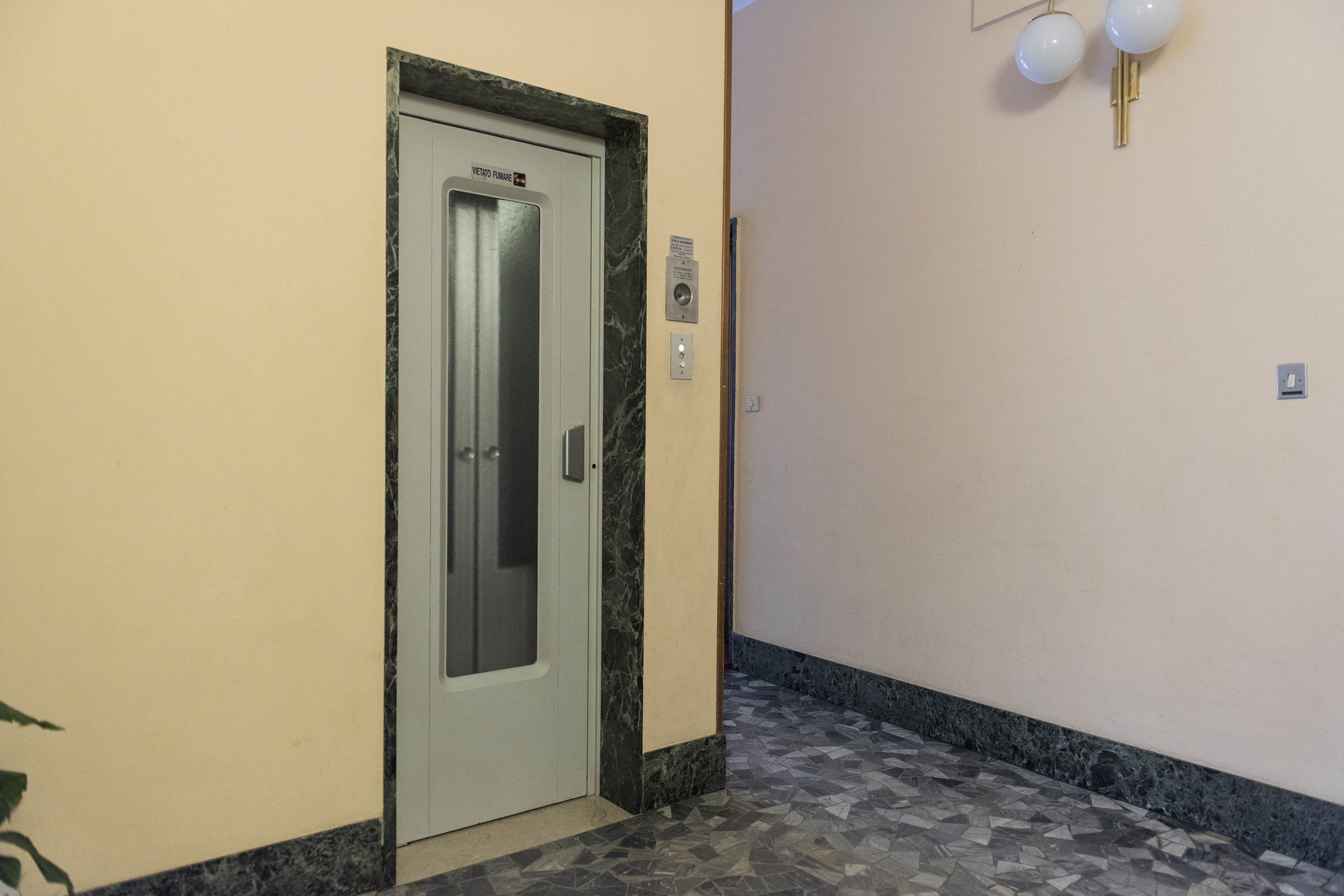An elevator door with a marble frame, a wall-mounted light fixture, and a patterned floor.