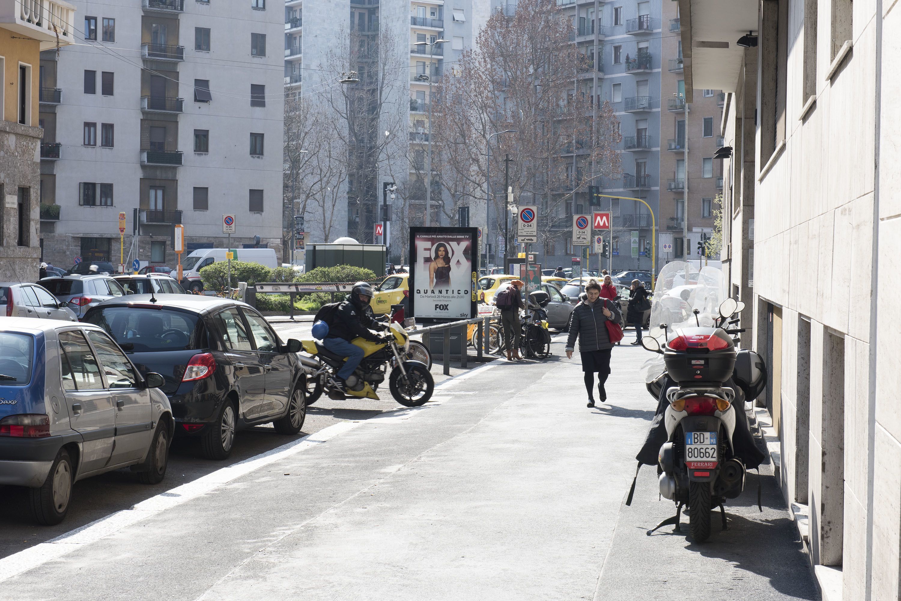 Een levendige stadstraat met geparkeerde auto's en motorfietsen, gebouwen op de achtergrond en een heldere lucht.