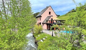 A traditional chalet with a wooden deck and a small balcony, surrounded by lush greenery and a flowing stream. - Traumhaftes Schwarzwaldhaus mit Swimmingpool (Freiburg im Breisgau)