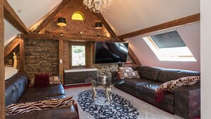 Un salón estilo loft con vigas de madera a la vista, pared de piedra, candelabro, televisor grande y una zona de estar acogedora.