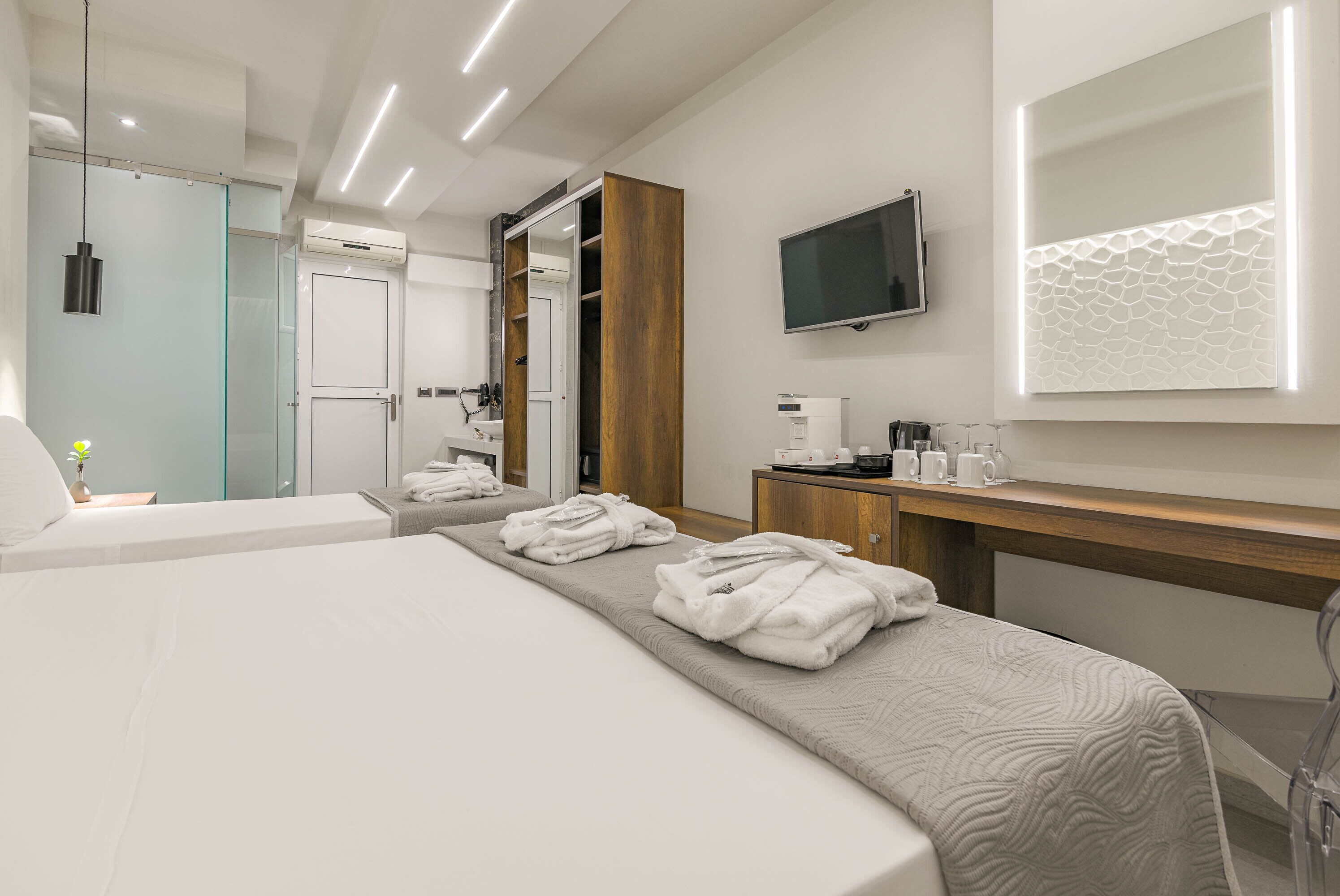 A modern hotel room with a large bed, a wooden wardrobe, a bedside table with a coffee maker, and a mounted TV.