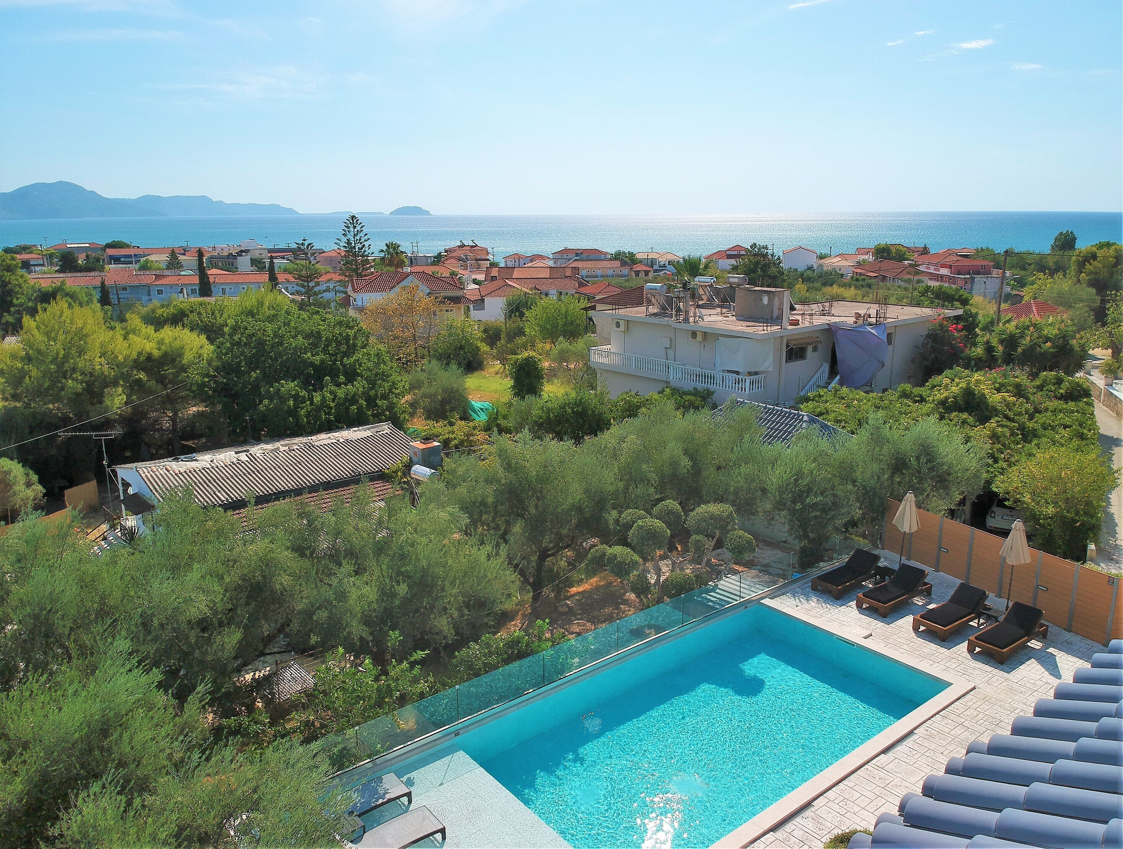 A rooftop pool with clear blue water, surrounded by lush greenery and overlooking a coastal town with red-roofed buildings.