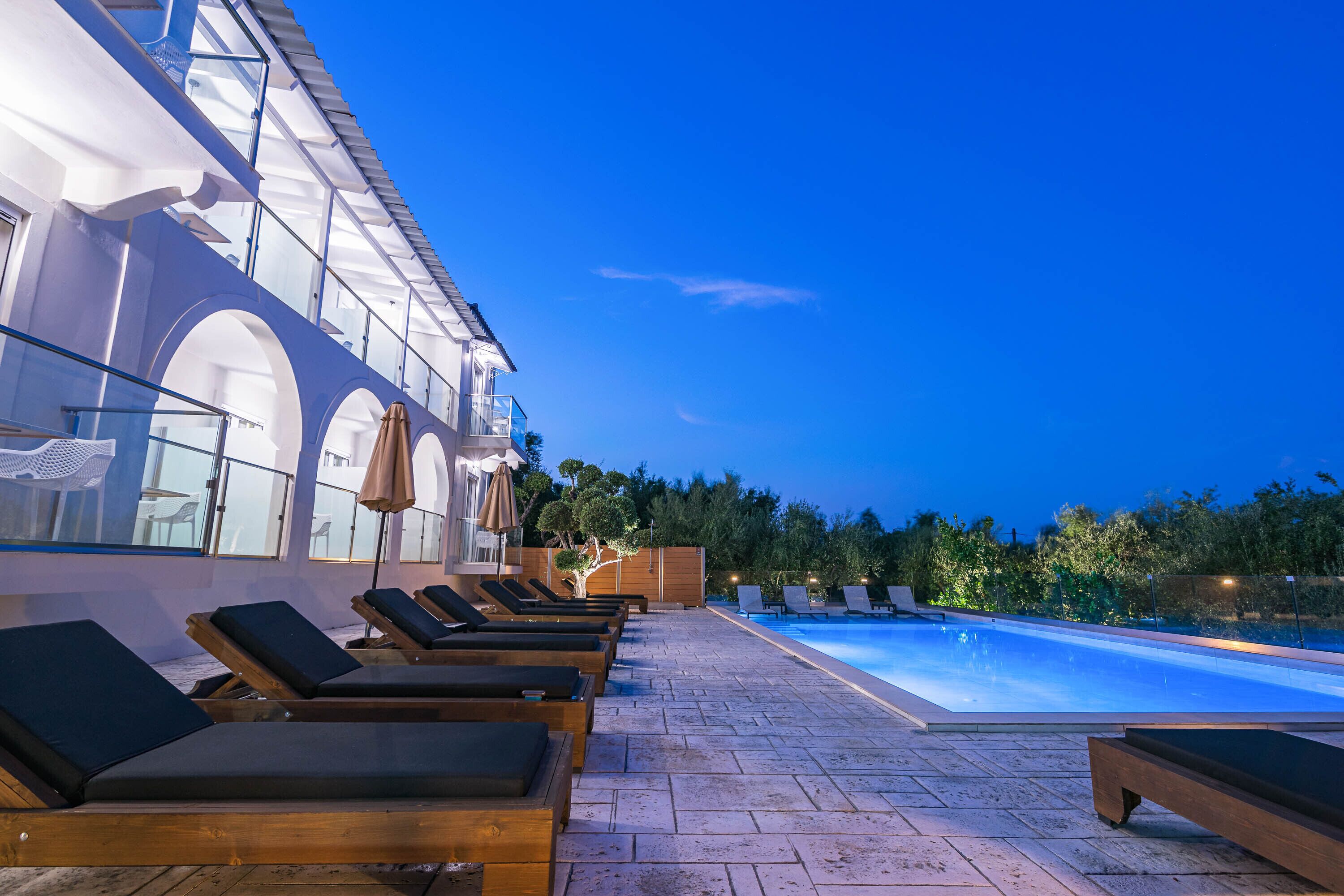 A poolside area with sun loungers and a swimming pool, surrounded by a building with large windows and a wooden deck.