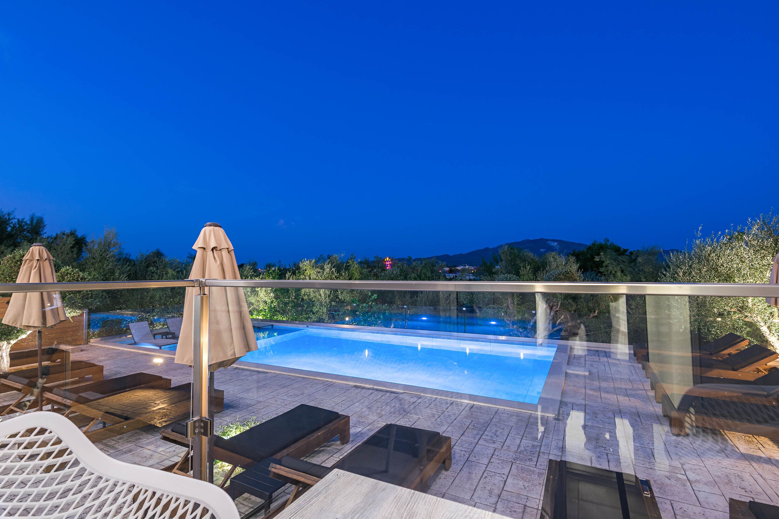A rooftop pool area with lounge chairs and umbrellas, surrounded by a glass railing.
