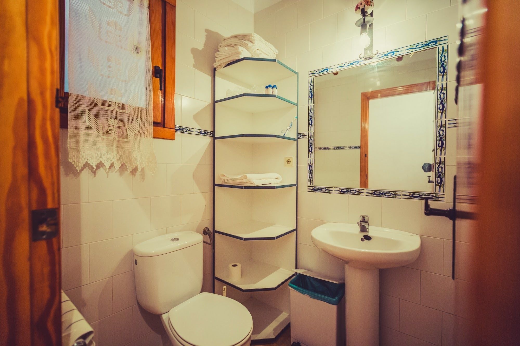A bathroom with a toilet, a sink, and a mirror. There are shelves above the toilet and a towel rack.