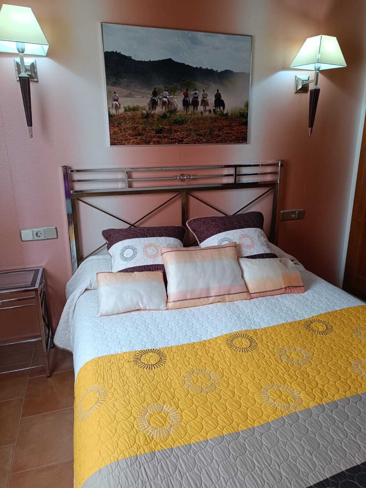 A neatly made bed with a yellow and gray quilt, two wall-mounted lamps, and a painting of a group of people on horseback above the bed.