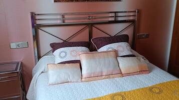 Una cama con ropa de cama bien tendida, con un colcha amarilla y gris, dos lámparas fijadas a la pared y un cuadro de un grupo de personas a caballo sobre la cama.