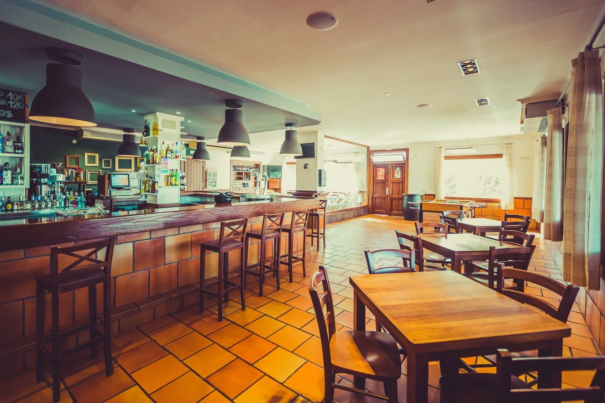 Un espace bar avec des tabourets et un comptoir en bois, un espace repas avec des tables et des chaises, et une cuisine visible au fond.