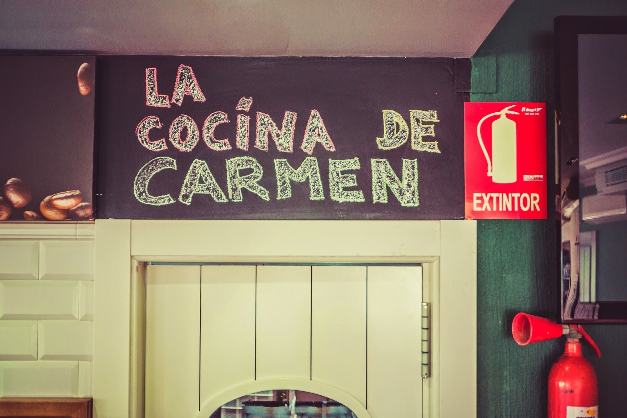A kitchen area with a chalkboard sign reading "LA COCINA DE CARMEN" and a red fire extinguisher mounted on the wall.