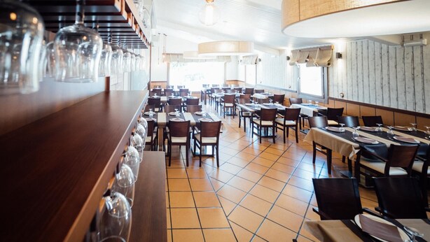 Un restaurante amplio con varias mesas y sillas, una barra alargada con copas de vino colgantes y una decoración de estilo moderno.