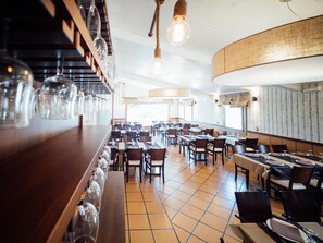 Un restaurante amplio con varias mesas y sillas, una barra alargada con copas de vino colgantes y una decoración de estilo moderno.