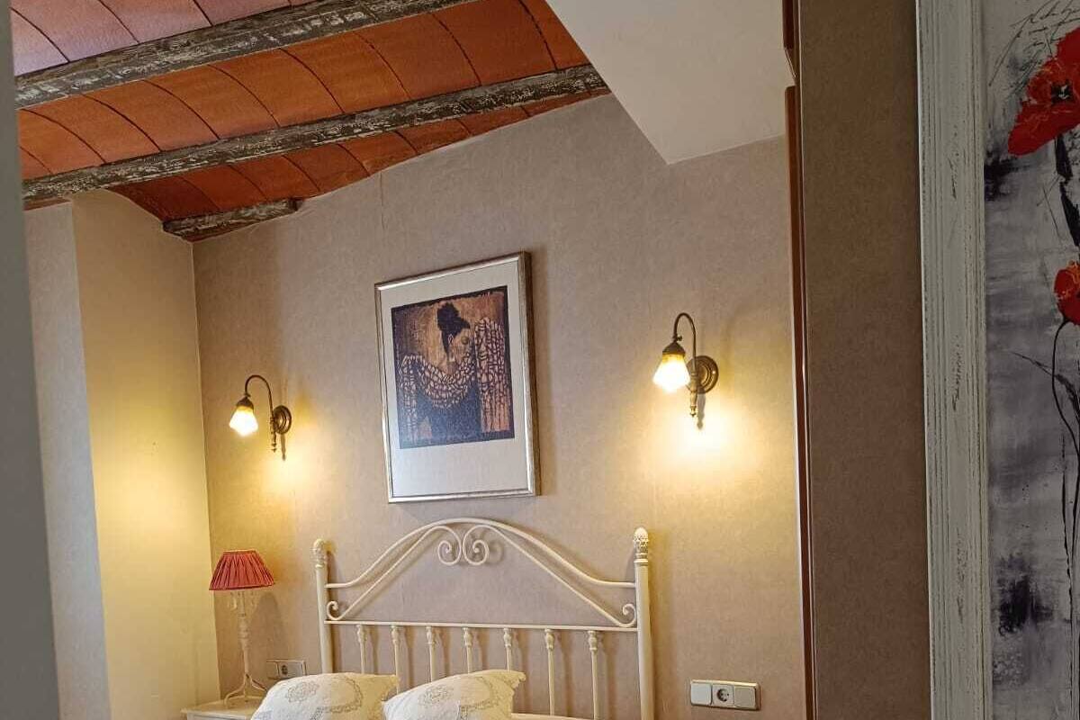 Een slaapkamer met een wit bed, twee wandlampen, een lijst met een kunstwerk erboven en een rode lamp op een nachtkastje.