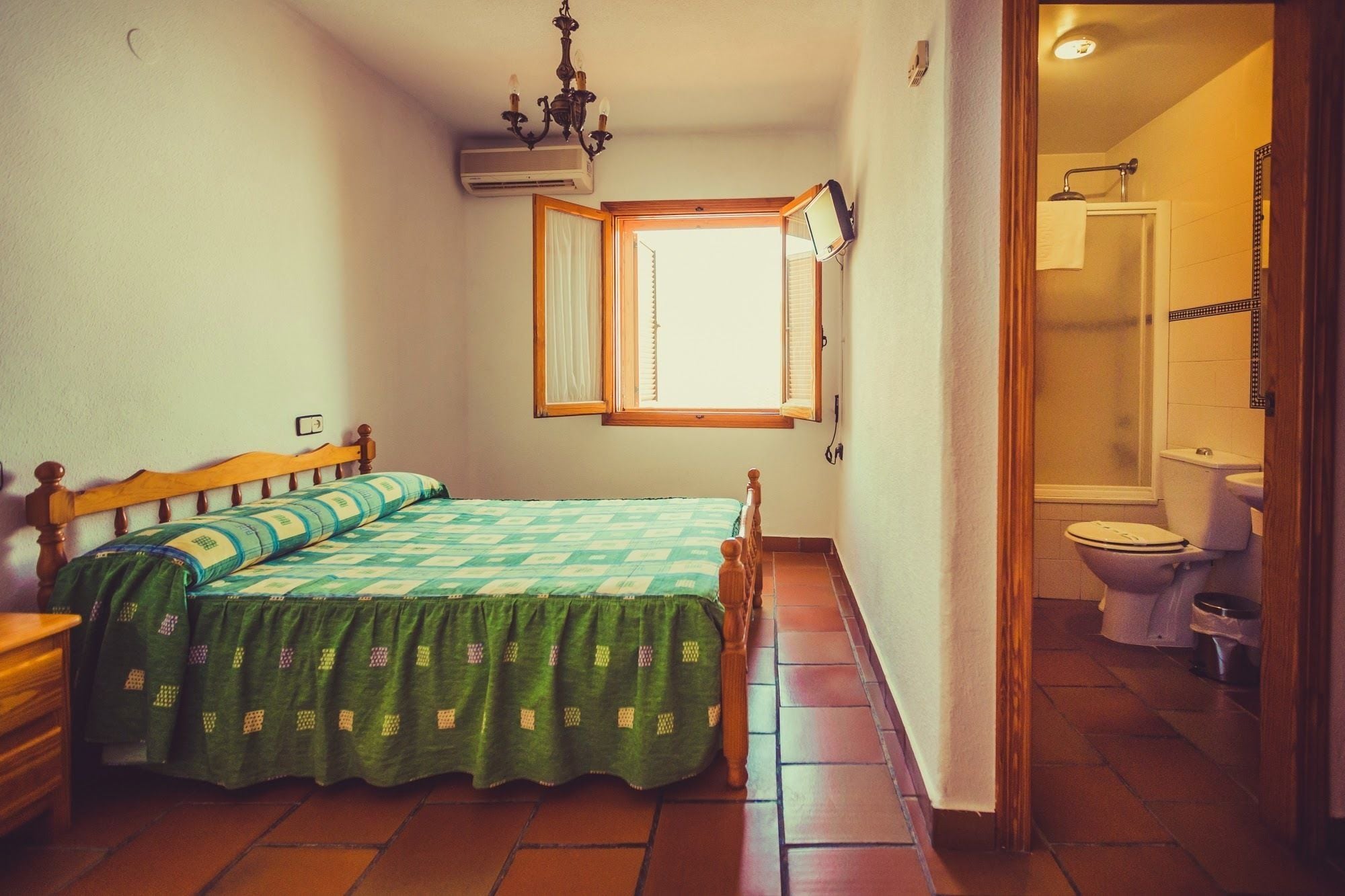 Un dormitorio pequeño y bien iluminado con una cama individual, un aparador de madera y una ventana con contraventanas abiertas.