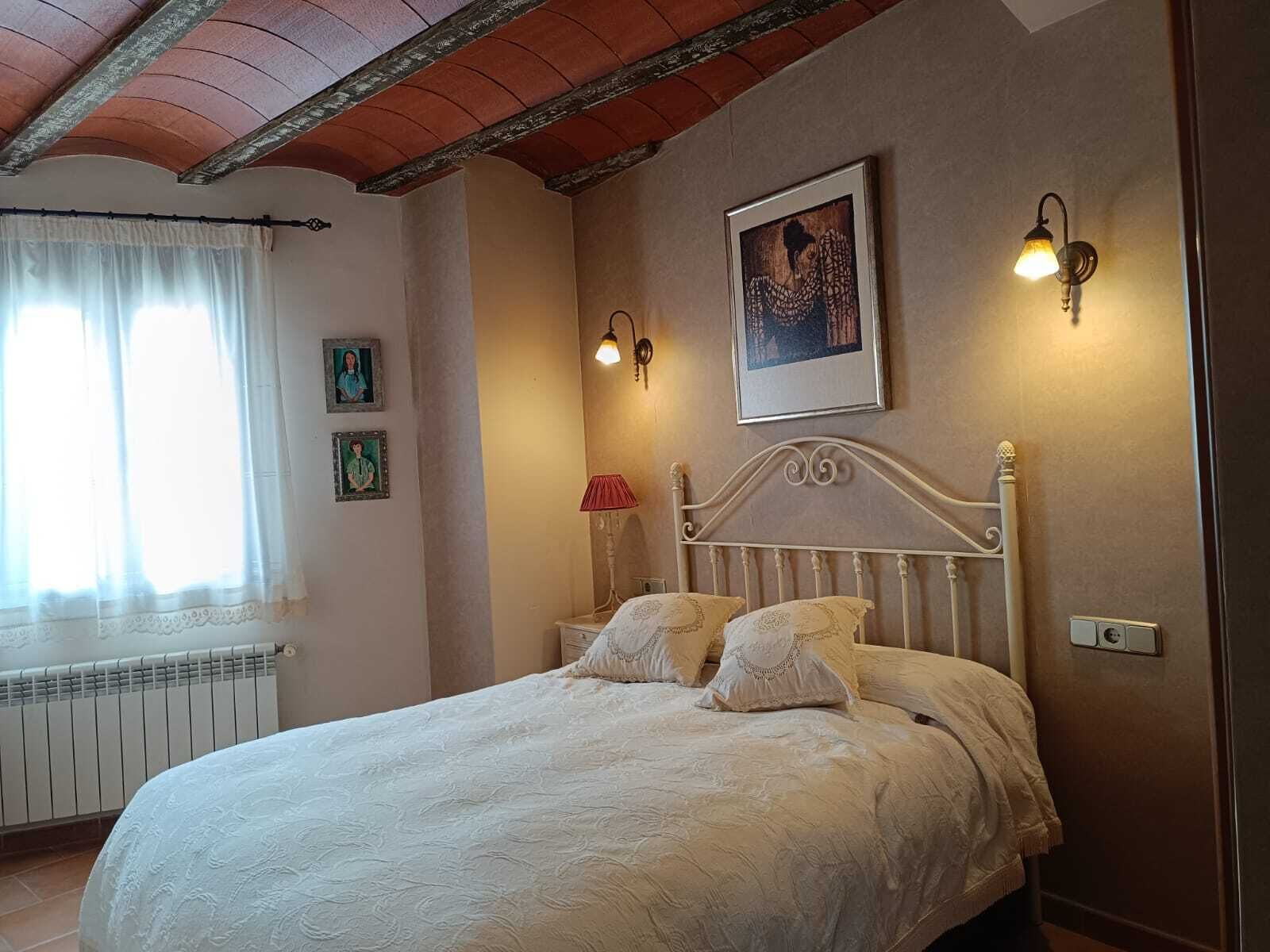 A bedroom with a bed, a window with curtains, a radiator, wall-mounted lights, and framed pictures.