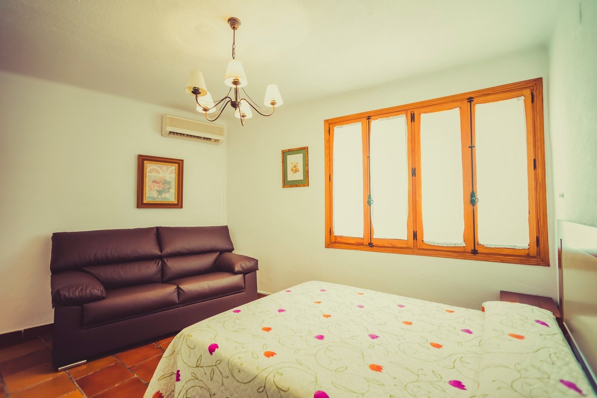 A bedroom with a brown leather sofa, a bed with a floral bedspread, a wooden-framed window, and a chandelier.