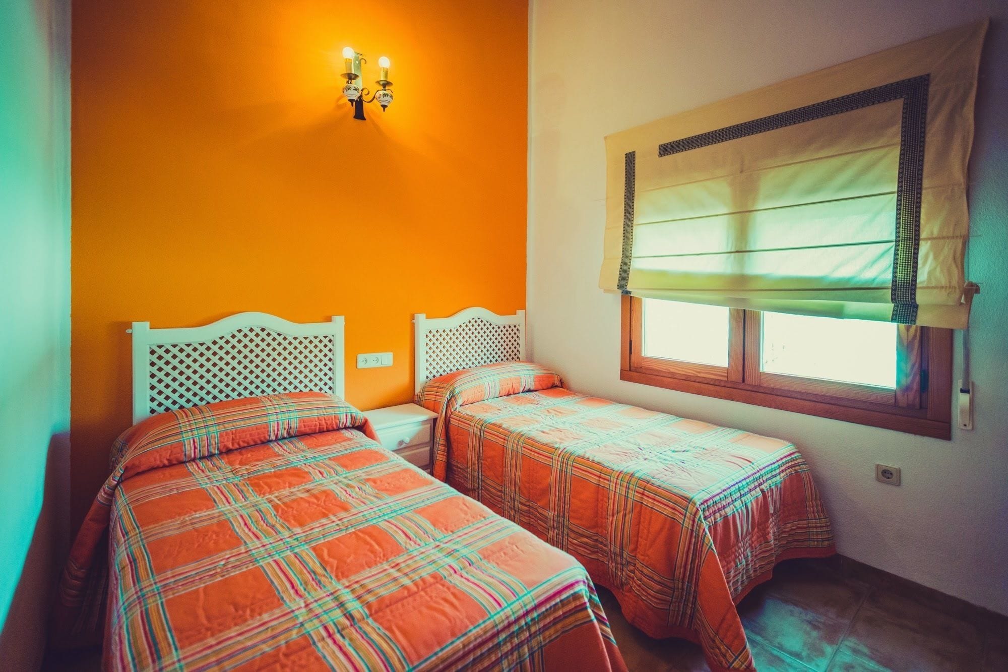 A room with two beds, a window with a curtain, and a wall-mounted light fixture.
