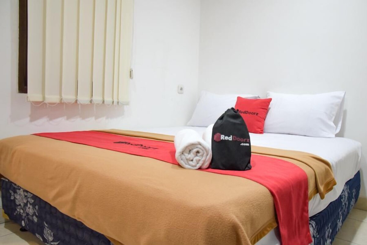 A neatly made bed with a red and beige bedspread, a rolled white towel, and a black bag with "Red Doorz" on it.