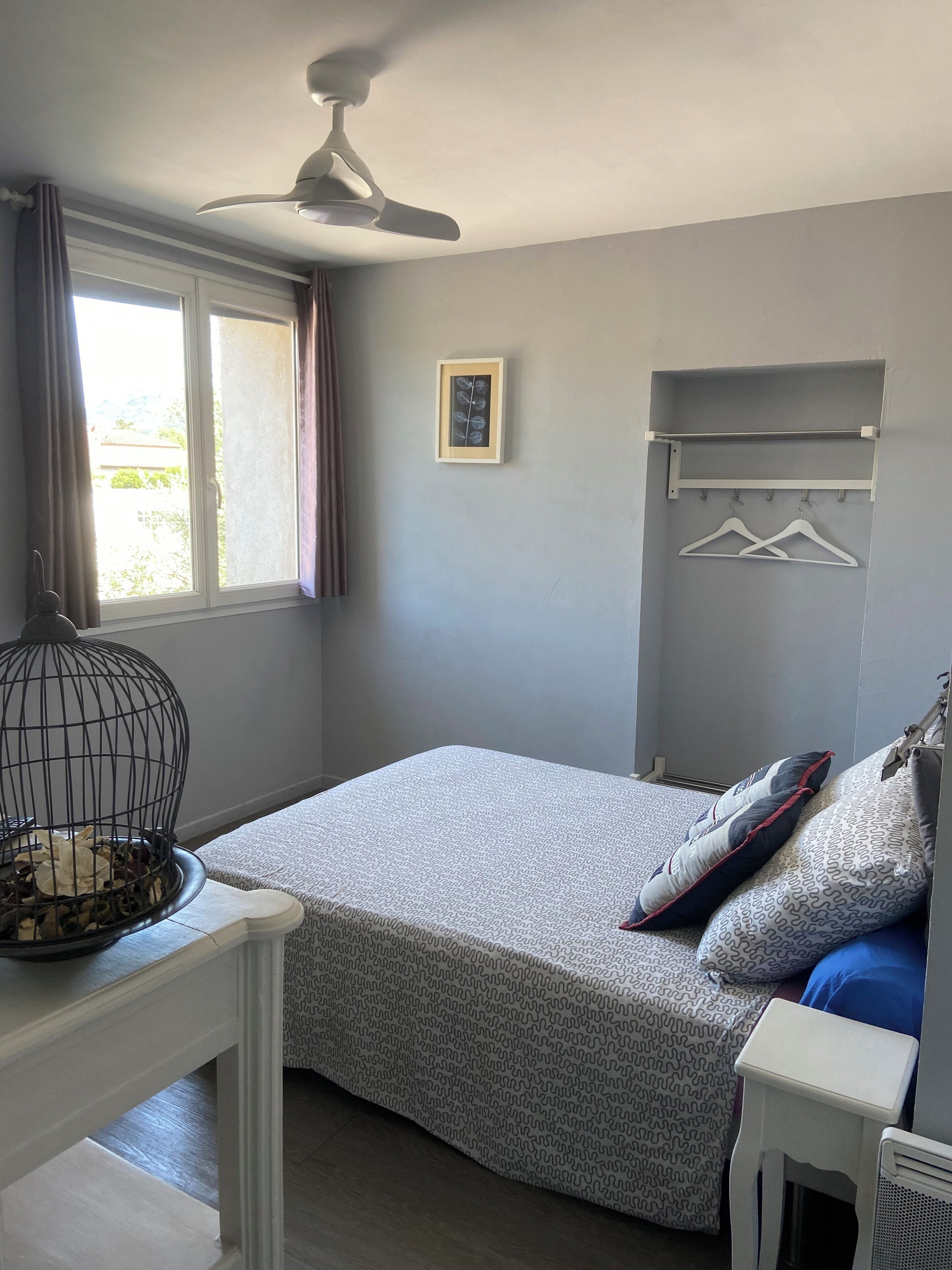 A bedroom with a bed, a ceiling fan, a window with curtains, a framed picture, and a coat rack with hangers.