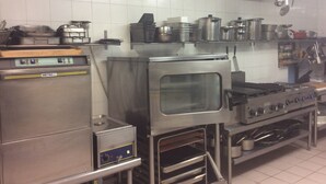 Una cocina comercial con electrodomésticos de acero inoxidable, incluyendo un horno grande y una estufa, además de varios utensilios y recipientes de cocina en estanterías.